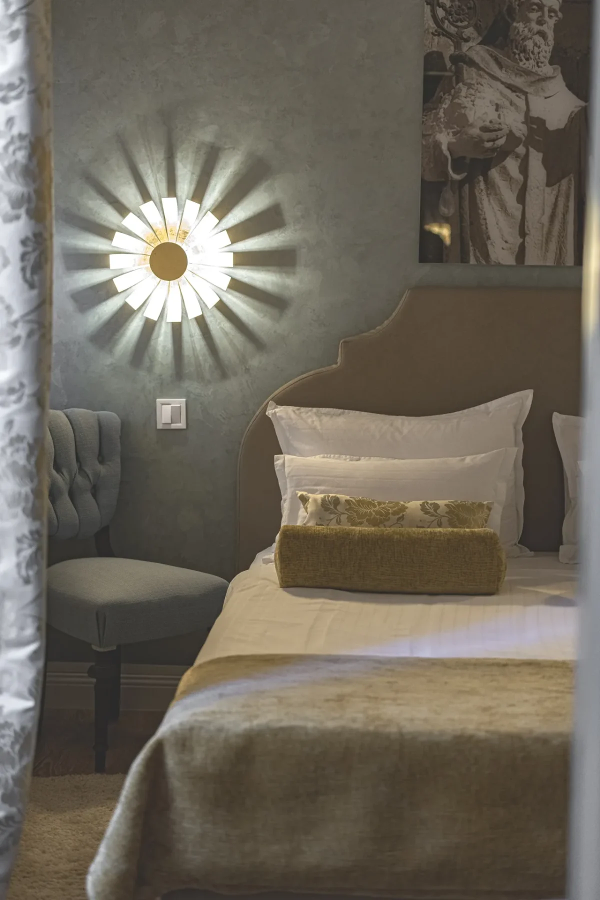 Double bed and gold sunburst wall light seen through softly draped curtains in the Deluxe Double Room