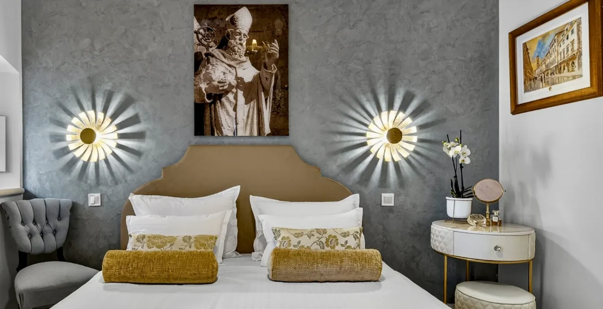 Upholstered headboard between two gold sunburst wall lamps and a print of St. Blaise, patron saint of Dubrovnik
