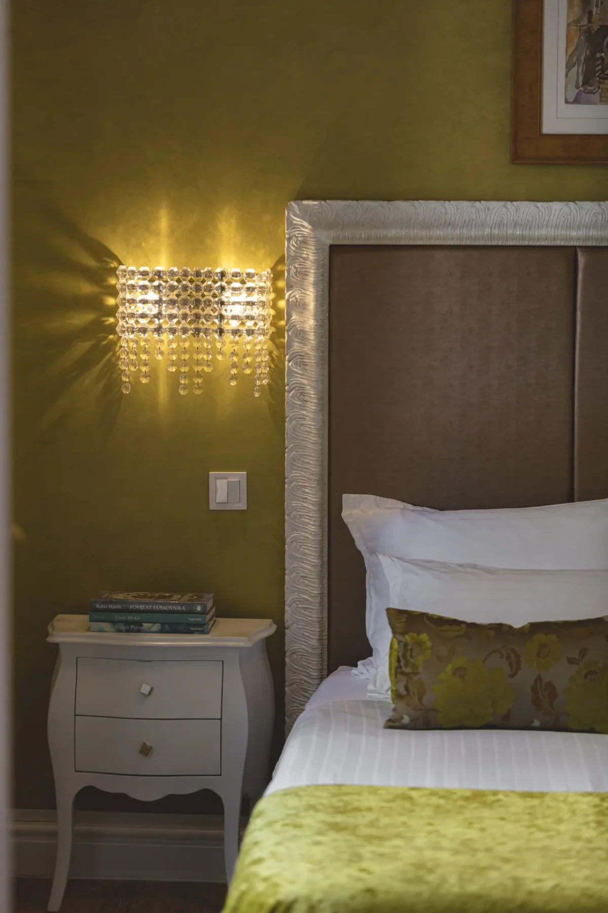 White bedside table with books next to the bed and crystal wall light in the Executive Suite