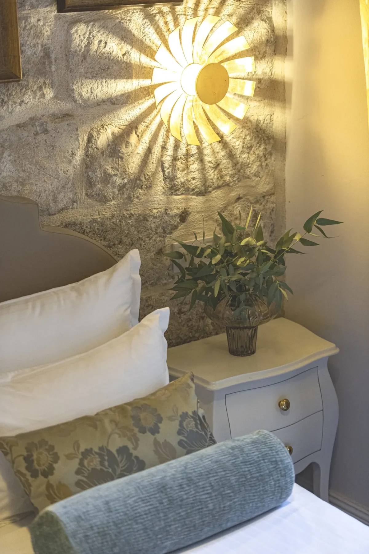 Warm-lit bedside corner with stone wall, potted greenery and sunburst wall lamp in the Premium Double Room